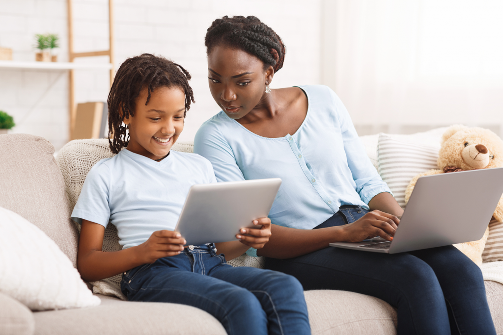 Woman working on laptop and sitting on couch next to a child looking at a tablet