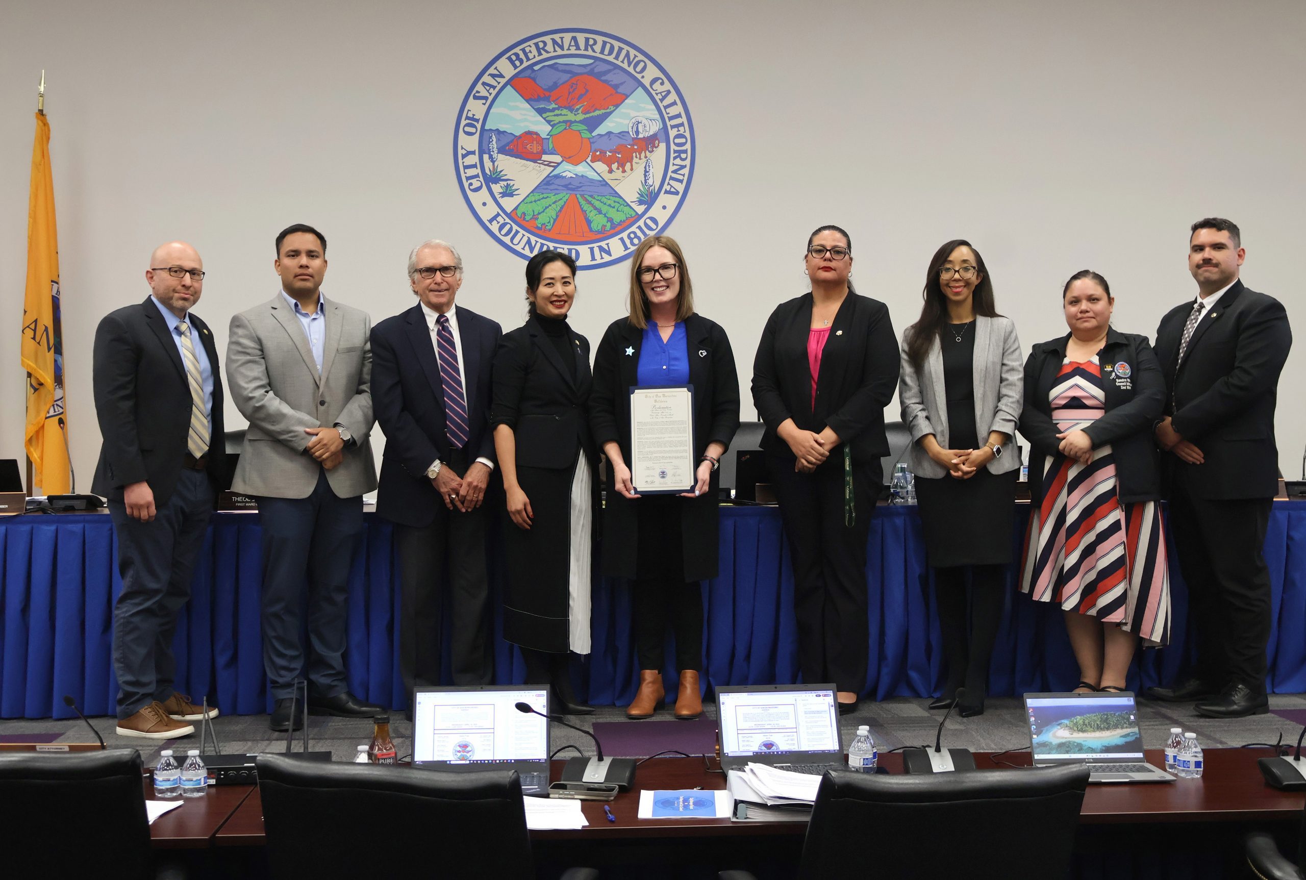 Group of nine people standing and smiling in a council room, while a woman holds a proclamation letter for child abuse prevention month.