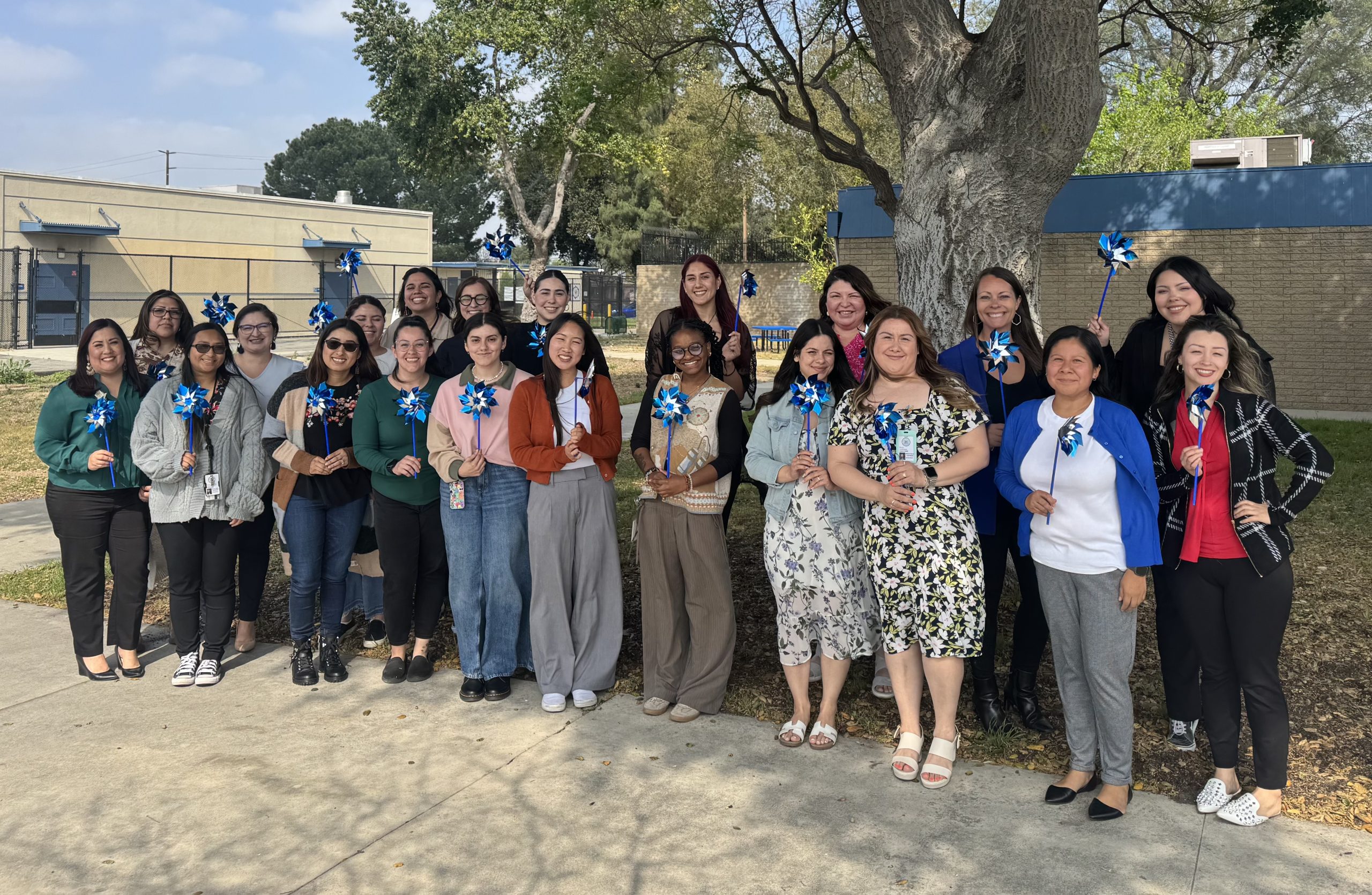 Large team of people stand outside holding blue and silver pinwheels together in front of a tree for Child Abuse Prevention Month.