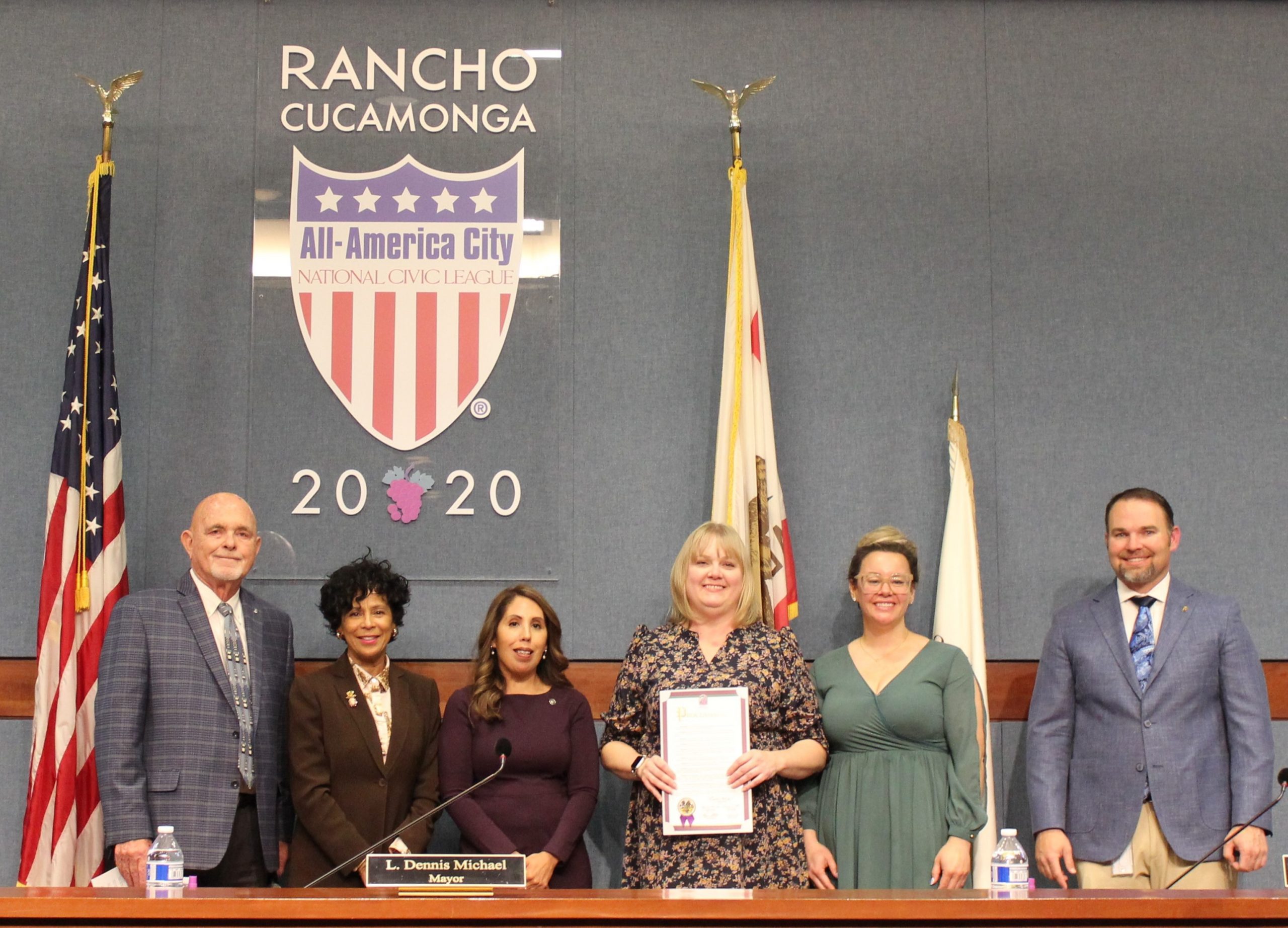 Group of six people standing and smiling in a council room, while a woman holds a proclamation letter for child abuse prevention month.
