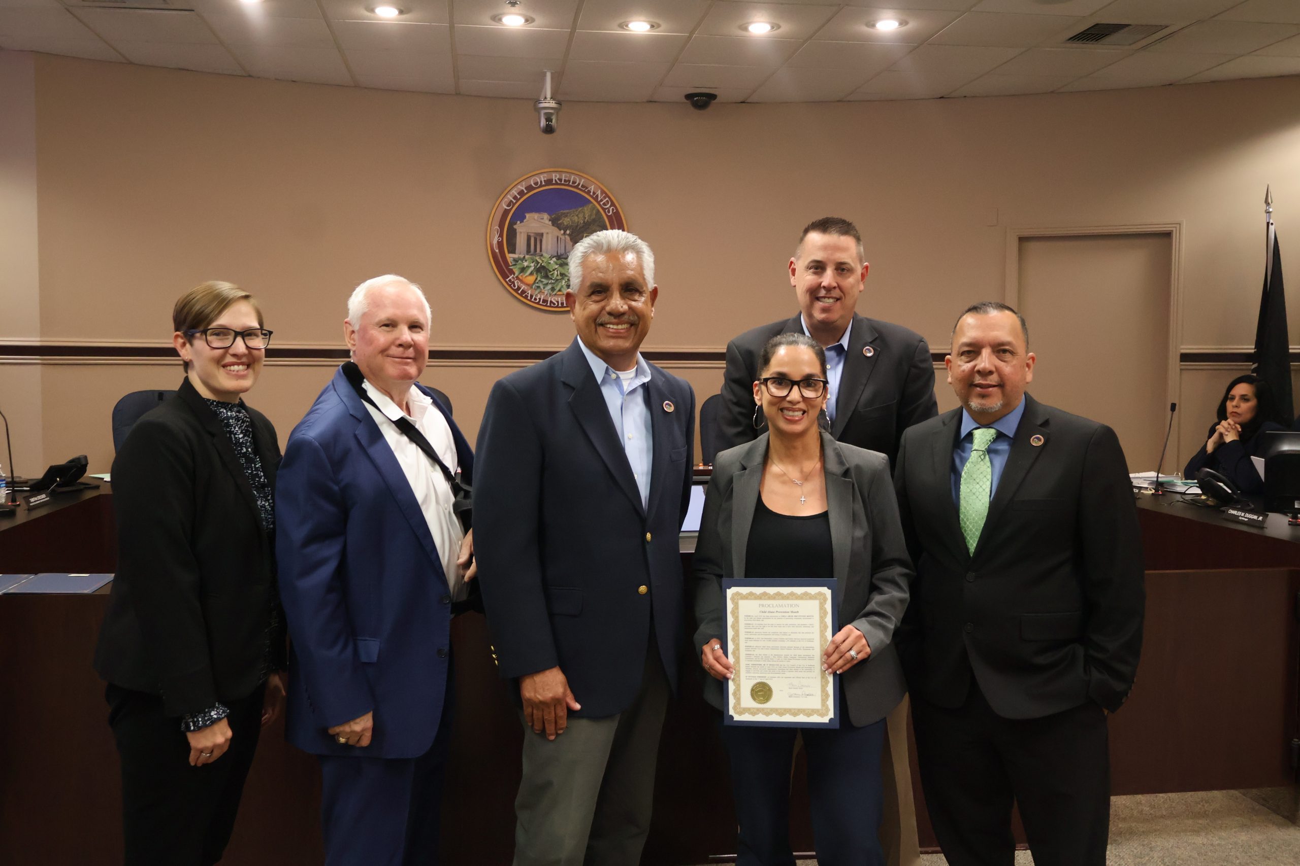 Group of six people standing and smiling in a council room, while a woman holds a proclamation letter for child abuse prevention month.