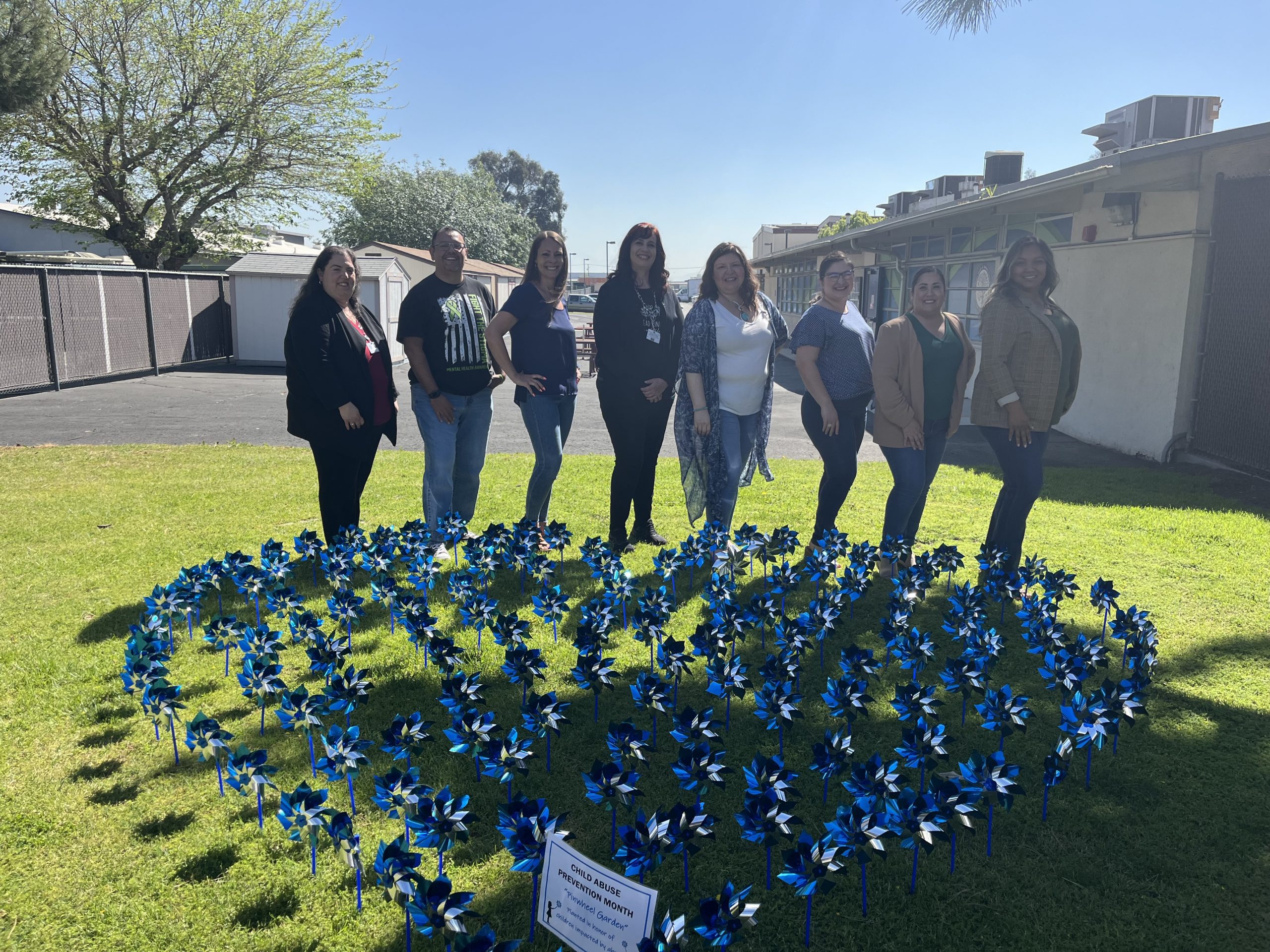 A smaller group of people stand behind a heart-shaped pinwheel garden on green grass.